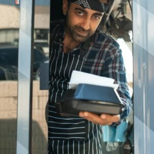 Male food vendor presenting a takeout box from a food truck window, wearing plaid shirt and apron.